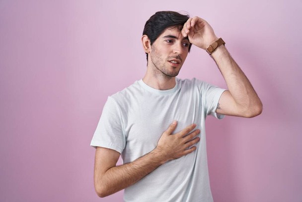 Jonge Spaanse man die over roze achtergrond staat en het voorhoofd aanraakt voor ziekte en koorts, griep en kou, virusziek  - Foto, afbeelding