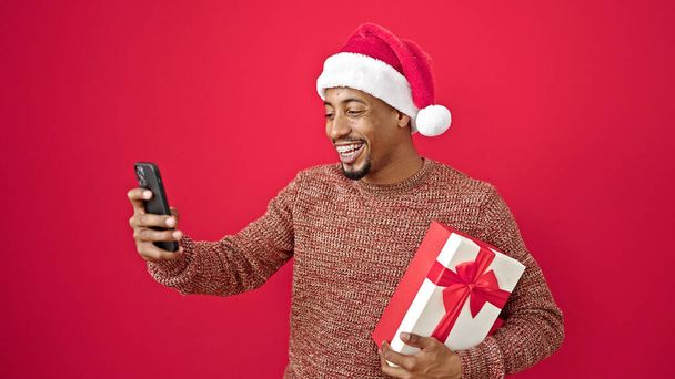 Afrikaans amerikaans man holding christmas gift het hebben van video oproep over geïsoleerde rode achtergrond - Foto, afbeelding