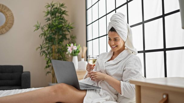 Jovem bela mulher hispânica vestindo roupão de banho bebendo champanhe tendo chamada de vídeo no quarto - Foto, Imagem