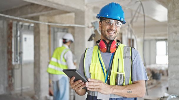 Dos hombres constructores sonriendo confiados usando touchpad en el sitio de construcción - Foto, Imagen
