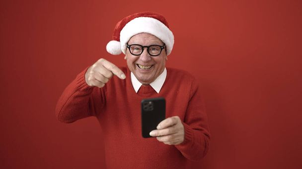 senior nosí vánoční klobouk ukazující na smartphone přes izolované červené pozadí - Fotografie, Obrázek