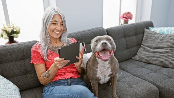 Αυτοπεποίθηση γκριζομάλλης μεσήλικας γυναίκα που χρησιμοποιεί την πιστωτική κάρτα στο touchpad για online αγορές, κάθεται με το σκυλί της στον καναπέ στο σπίτι - Φωτογραφία, εικόνα