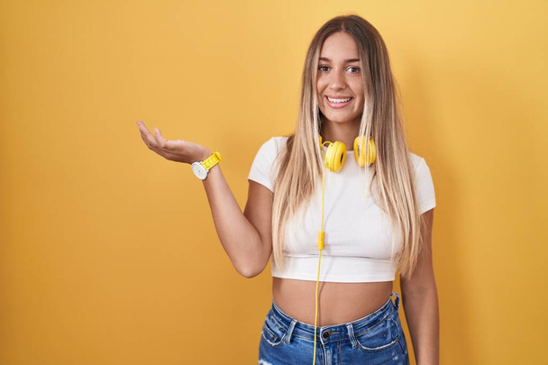 Jonge blonde vrouw die over een gele achtergrond staat met een koptelefoon in haar glimlachende, vrolijke presentatie en wijzend met handpalm naar de camera kijkend.  - Foto, afbeelding