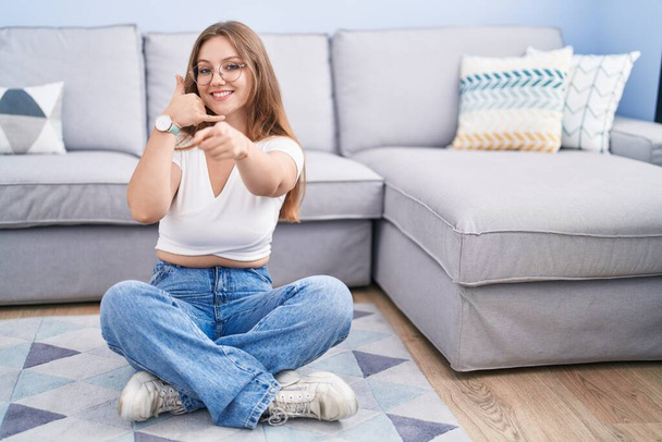 Young caucasian woman sitting on the floor at the living room smiling doing talking on the telephone gesture and pointing to you. call me.  - Photo, Image