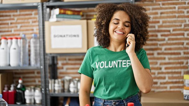 Fiatal gyönyörű spanyol nő önkéntes beszél okostelefonon mosolyog jótékonysági központ - Fotó, kép