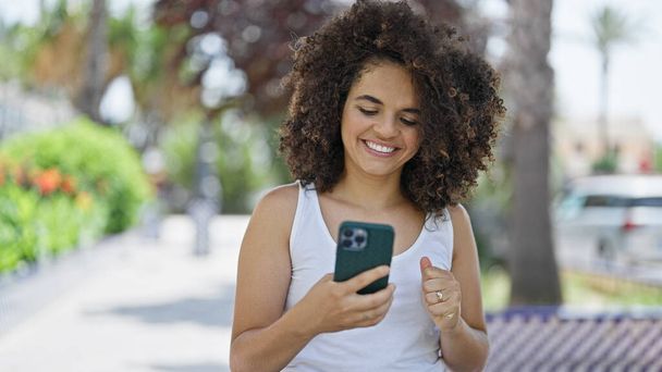 Молода красива іспаномовна жінка використовує смартфон, посміхаючись у парку
 - Фото, зображення