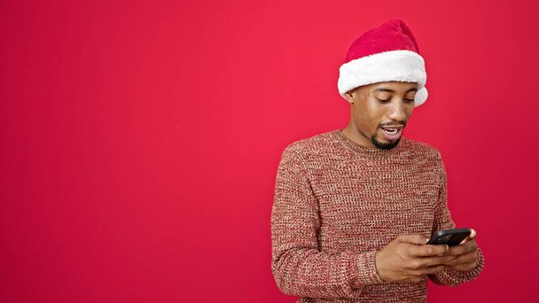 Afrikaanse amerikaanse man het dragen van kerst hoed met behulp van smartphone glimlachen over geïsoleerde rode achtergrond - Foto, afbeelding