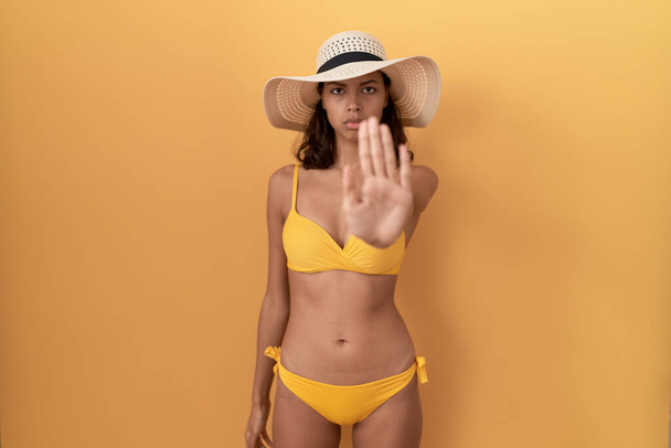 Jeune femme hispanique portant un bikini et chapeau d'été faire arrêter chanter avec la paume de la main. expression d'avertissement avec geste négatif et sérieux sur le visage.  - Photo, image
