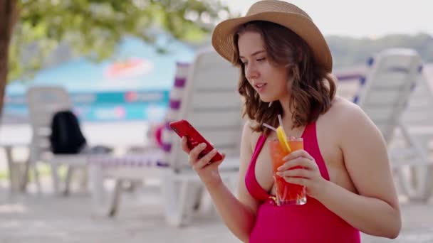 休暇中にソーシャルメディアを閲覧するボディポジティブガール. 赤いビキニの女性は,屋外のプールサイドで夏休みを投稿します. 元気な女性は,飲酒時に携帯電話でブログを書くことで水着を着た - 映像、動画
