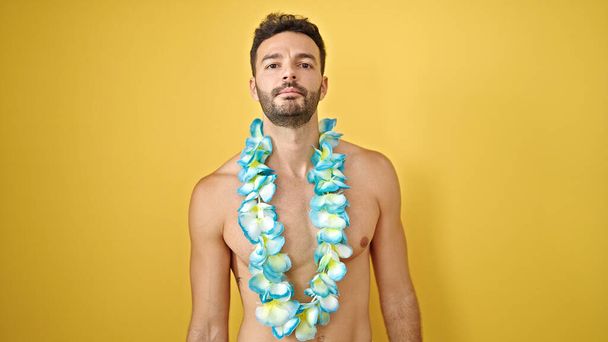 Junger hispanischer Tourist in hawaiianischem Lei steht ohne Hemd mit ernstem Gesicht vor isoliertem gelben Hintergrund - Foto, Bild