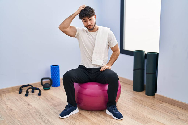 Hispanic man met baard zittend op pilates ballen in yoga kamer verwarren en vragen stellen over de vraag. onzeker met twijfel, denken met de hand op het hoofd. duur concept.  - Foto, afbeelding