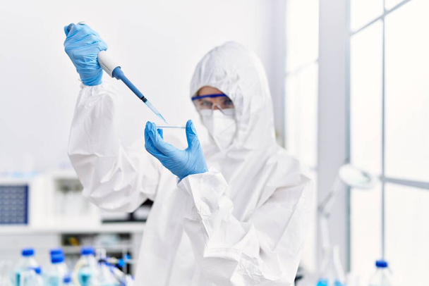 Junge blonde Wissenschaftlerin in Sicherheitsuniform gießt Flüssigkeit auf Probe im Labor - Foto, Bild