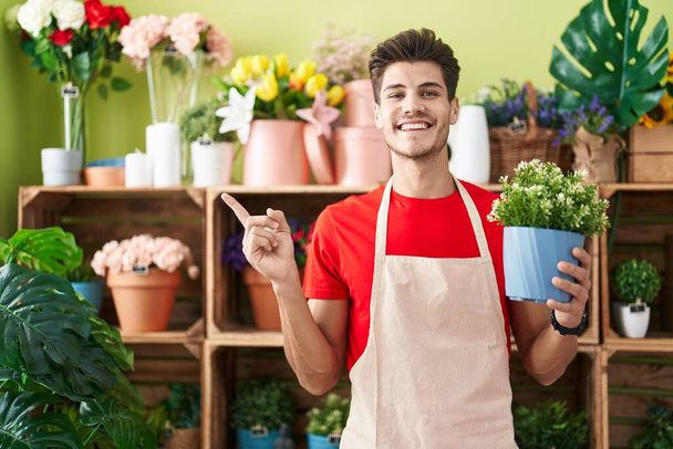 Młody Hiszpan pracujący w kwiaciarni, trzymający roślinę uśmiechniętą z uśmiechem na dłoni i palcu.  - Zdjęcie, obraz