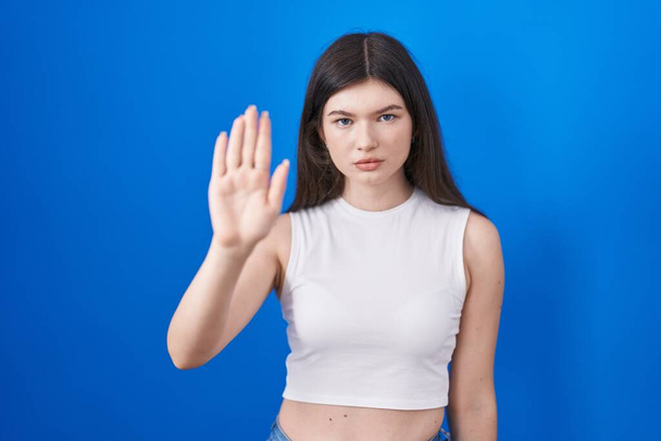 Jonge blanke vrouw die over een blauwe achtergrond staat te zingen stop met handpalm van de hand. waarschuwingsuitdrukking met negatief en ernstig gebaar op het gezicht.  - Foto, afbeelding