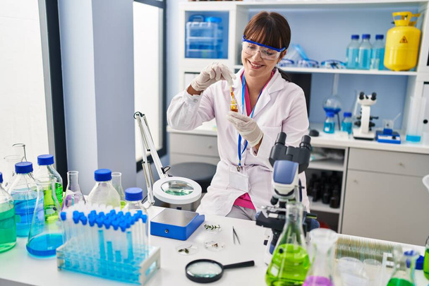Young beautiful hispanic woman scientist holding bottle with liquid at pharmacy - Photo, Image