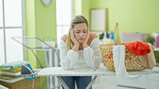 Young blonde woman leaning on ironing table with sad expression at laundry room - Photo, Image