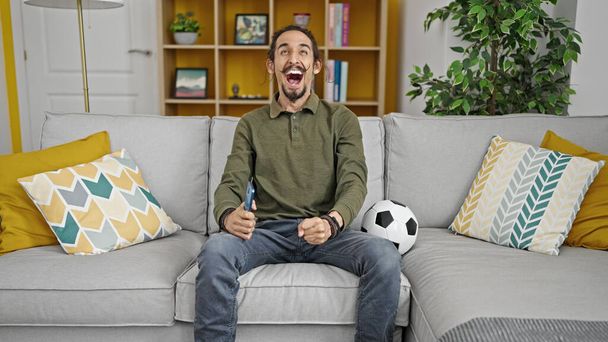 Junger hispanischer Mann unterstützt Fußballmannschaft sitzt zu Hause auf Sofa - Foto, Bild