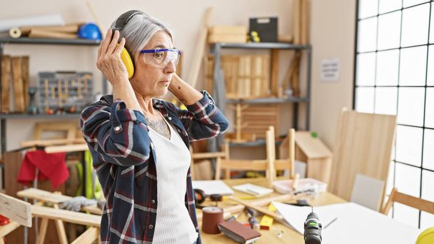 Serious middle age grey-haired woman carpenter working industriously, donned in safety headphones and security glasses at her indoor carpentry workshop. - Photo, Image