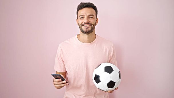 孤立したピンクの背景の上にサッカーボールを保持するスマートフォンを使用して若いヒスパニック男性 - 写真・画像