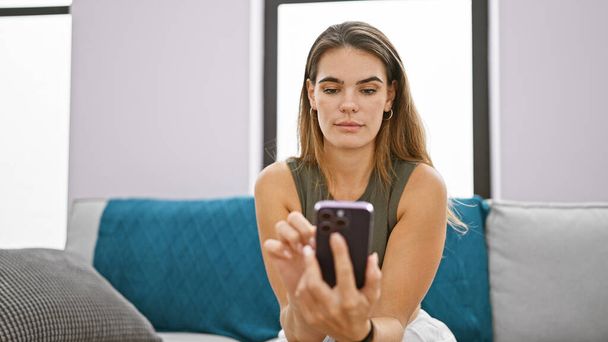 Fiatal gyönyörű spanyol nő használja okostelefon ül kanapén gondolkodás otthon - Fotó, kép