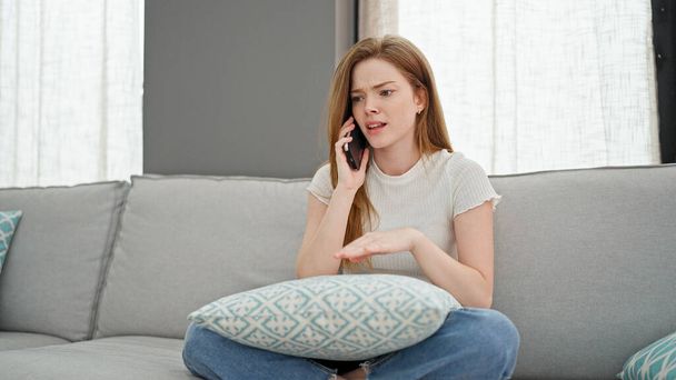 Mujer rubia joven hablando en el teléfono inteligente sentado en el sofá discutiendo en casa - Foto, Imagen