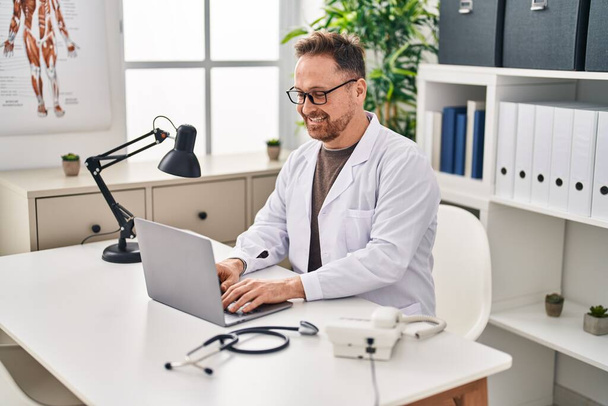 Klinikte çalışan genç beyaz bir doktor dizüstü bilgisayar kullanıyor. - Fotoğraf, Görsel