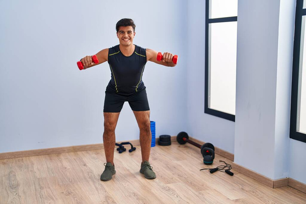 Молодий іспаномовний чоловік посміхається впевнено використовуючи тренування гантелів у спортивному центрі
 - Фото, зображення