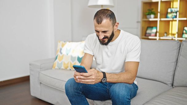 Giovane uomo ispanico utilizzando smartphone seduto sul divano a casa - Foto, immagini