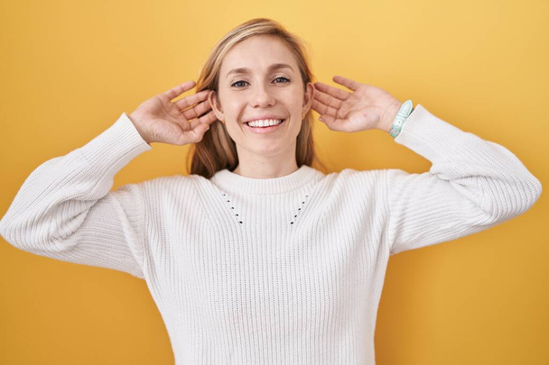 Jeune femme caucasienne portant un pull blanc sur fond jaune souriant tirant les oreilles avec les doigts, geste drôle. problème d'audition  - Photo, image