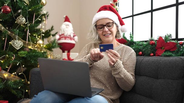 Женщина средних лет с седыми волосами делает рождественские покупки онлайн с ноутбуком дома - Фото, изображение