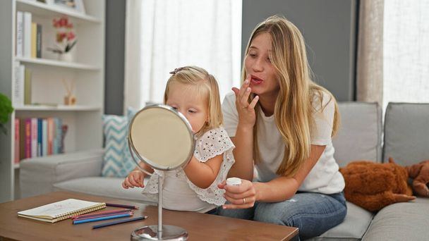 Relaxed caucasian mother and daughter cosy at home, lying comfortably on sofa, seriously applying makeup on lips, looking into mirror - Photo, Image