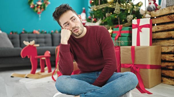 Young hispanic man celebrating christmas sitting on floor looking upset at home - Photo, Image
