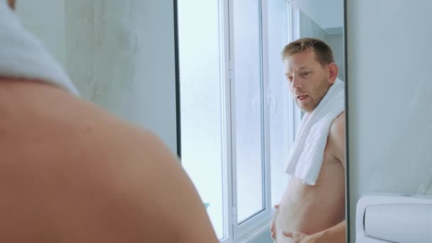 Fiatal, elégedett fehér férfi törölközőben, aki tükörben nézi a testét a fürdőszobában. Sovány elégedett férfi ellenőrzi a gyomor és a testzsír százalékos reggel. - Felvétel, videó