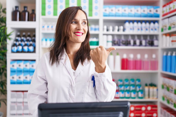 Femme brune d'âge moyen travaillant à la pharmacie drugstore souriant avec le visage heureux regardant et pointant vers le côté avec le pouce vers le haut.  - Photo, image