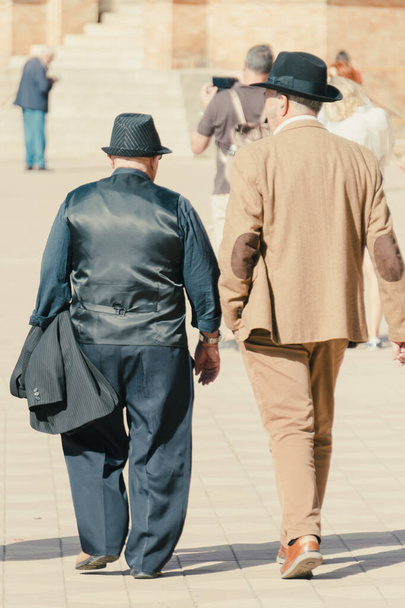 セビリア,アンダルシア,スペイン;11月16日2023:晴れた日にセビリアのプラザ・デ・パニャを歩くスーツと帽子を着た2人のエレガントな男性. - 写真・画像