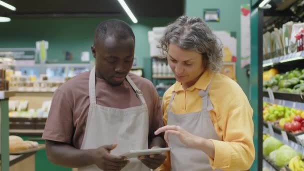 Portrait moyen de deux travailleurs de supermarchés masculins et féminins dans des tabliers regardant la tablette numérique et ayant la conversation puis souriant à la caméra debout ensemble dans l'allée avec l'épicerie - Séquence, vidéo