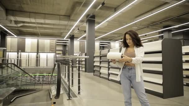 Eine junge Frau wählt beim Einkaufen in der Bauabteilung eines Verbrauchermarktes Tapeten für die Renovierung ihres Hauses. - Filmmaterial, Video