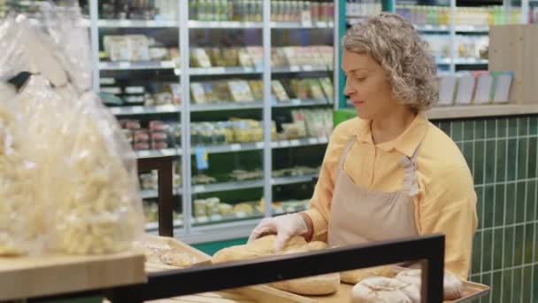 Tiro médio de funcionário de padaria feminino caucasiano que expõe pães frescos e baguetes em prateleiras de manhã - Filmagem, Vídeo