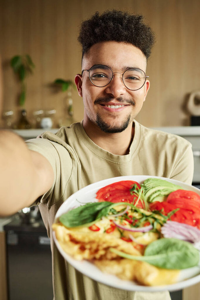 Headshot van lachende jonge vent uit het Midden-Oosten neemt foto van zichzelf met een bord met omelet en groenten - Foto, afbeelding
