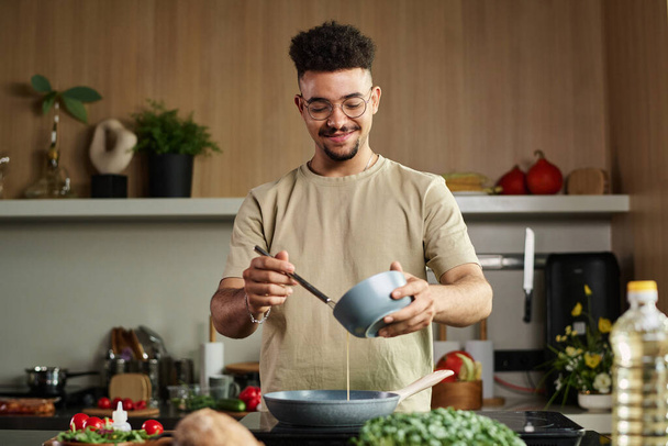 Средний снимок молодого парня с Ближнего Востока, который наливает взбитые яйца на сковороду, делая омлет, позируя у кухонной плиты. - Фото, изображение