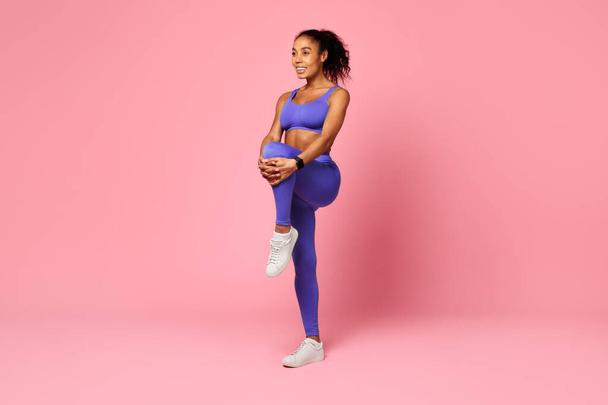 Trening i Wellness. Uśmiechnięta afro-amerykanka w sportowej odzieży przygotowująca się do treningu wytrzymałościowego, rozciągająca mięśnie nóg, trzymająca kolano przy klatce piersiowej nad różowym tłem studia. Pełna długość - Zdjęcie, obraz
