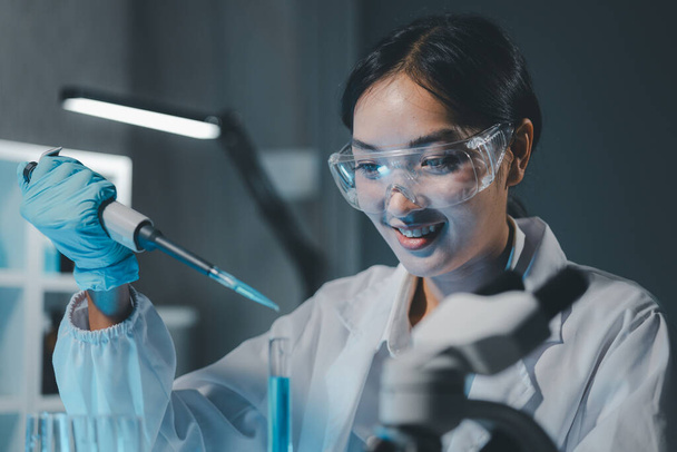 Fiatal tudósok steril ruházati és biztonsági szemüvegben, akik az asztalnál ülve kutatásokat végeznek egy orvosi laboratóriumban, mikroszkóppal, komoly koncentrált női mikrobiológussal.. - Fotó, kép