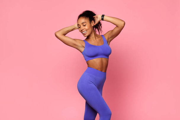 Fitness-Workout. Fit schwarze Frau trägt Sportbekleidung vor rosa Studiokulisse, hebt die Arme, hält die Hände hinter dem Kopf und genießt ihren perfekten schlanken Körper nach dem Training - Foto, Bild