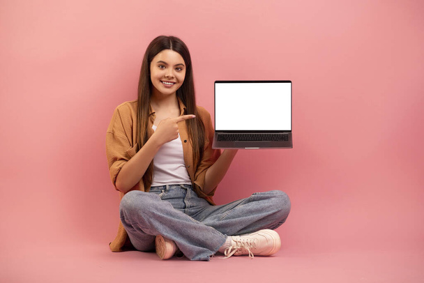 Great Website. Cheerful teen girl pointing at laptop with blank screen on her laps, happy female teenager demonstrating computer while sitting on floor over pink studio background, copy space, mockup - Photo, Image