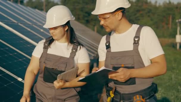 Zonnepark. Eco-engineering. Twee vrolijke mannelijke en vrouwelijke mechanische arbeiders lopen op landbouwgrond met zonne-energie-installatie verkennen. Samenwerkend team. Hoge kwaliteit 4k beeldmateriaal - Video