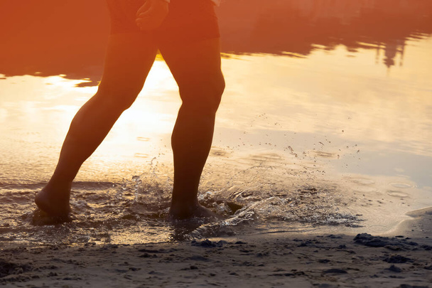 Una persona corre a lo largo de la playa en el agua, el mar o el océano en los rayos del sol, un atleta pasa tiempo felizmente, está entrenando, trotando al aire libre, vista de cerca de las piernas y salpicaduras. - Foto, imagen