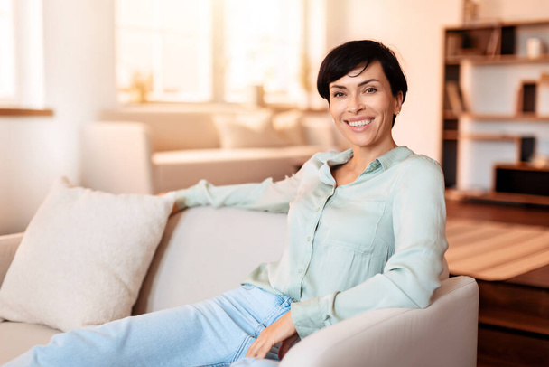 Radosna kobieta w średnim wieku uśmiecha się patrząc w kamerę, siedząc elegancko na sofie w salonie, ucieleśniając relaksujący weekend w domu. Wolne miejsce na tekst - Zdjęcie, obraz
