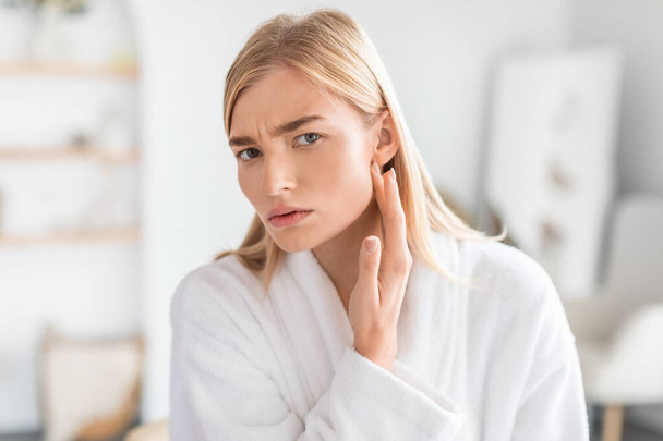 Young blonde woman examines her face skin closely for blackheads and wrinkles, showing worry in her modern bathroom at home. Skincare challenges and cosmetic treatment - Photo, Image