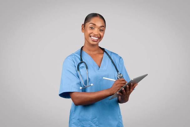Sonriente mujer milenaria negra doctora cirujana, enfermera en uniforme azul, toma notas en el portapapeles, aislada sobre fondo gris del estudio. Anuncio y oferta médica, examen, atención médica, ayuda profesional - Foto, imagen