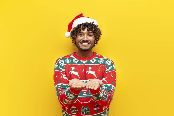 молодий кучерявий індуїст в новорічному одязі благає і дає порожні руки на жовтому ізольованому фоні, індуїстський хлопець в капелюсі Санта і різдвяний светр просить допомоги - Фото, зображення